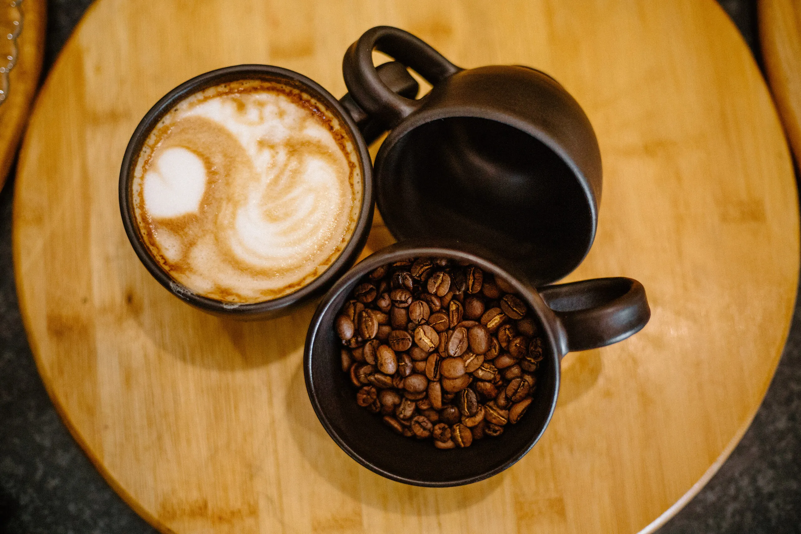 A cappuccino with latte art beside a bowl of roasted coffee beans on a wooden board